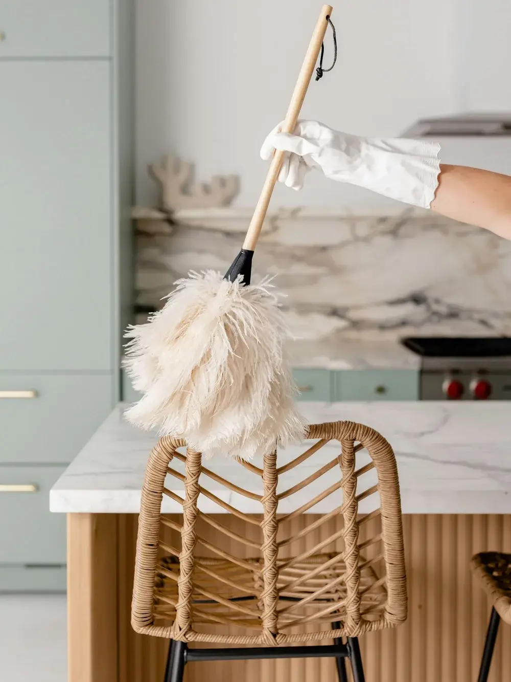 A white-gloved hand holding a feather duster above a rattan stool in a bright kitchen with a marble backsplash and counter