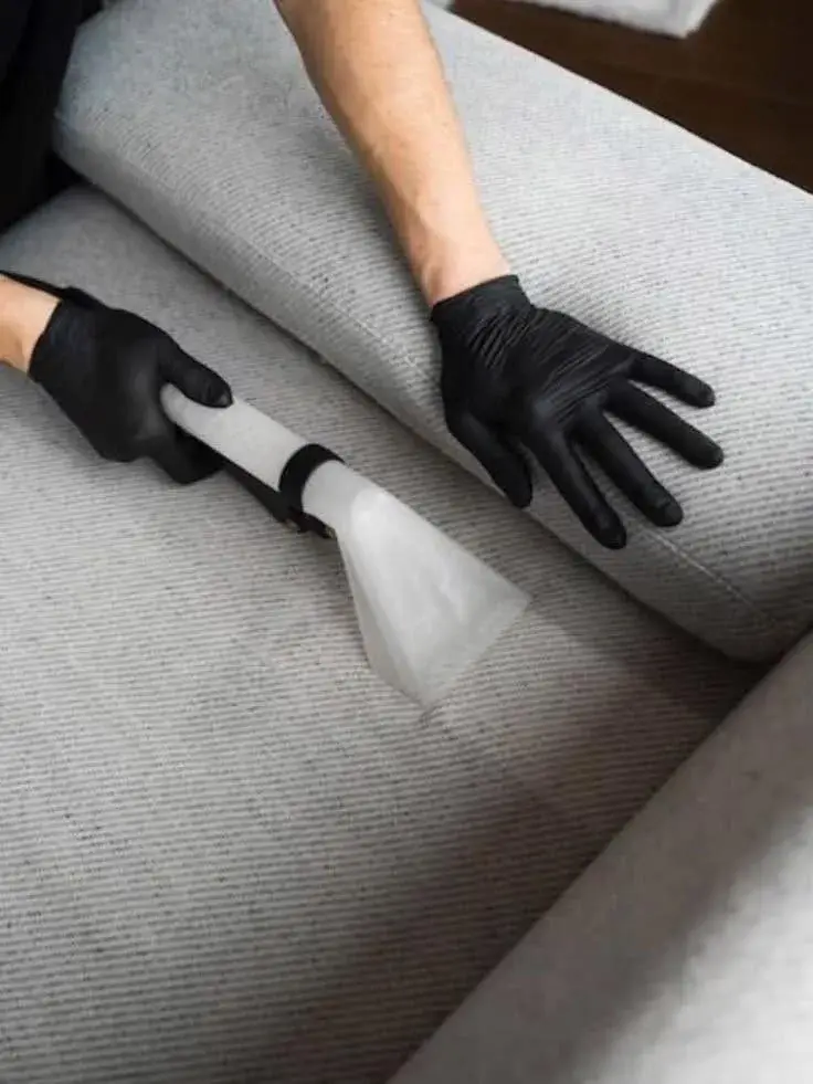 Black-gloved hands steam-cleaning the cushion of a grey fabric sofa